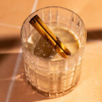 A glass of creamy white russian with a cinnamon stick is on a sunlit tile surface.