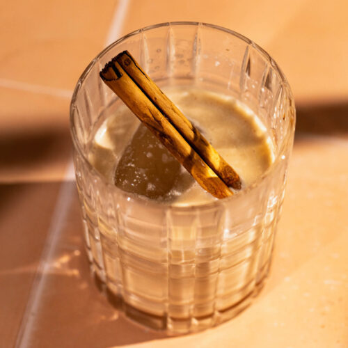 A glass of creamy white russian with a cinnamon stick is on a sunlit tile surface.