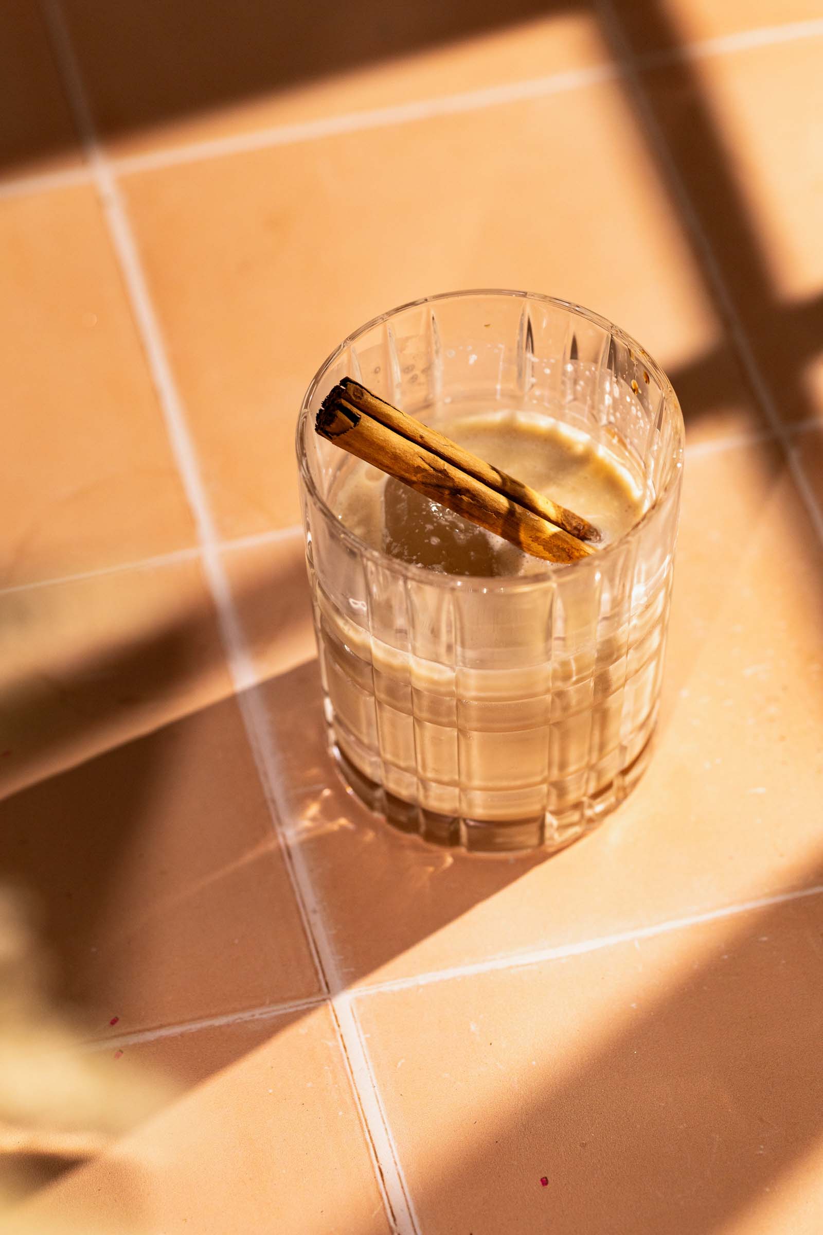 A creamy white russian topped with a cinnamon stick sits on a tiled surface in warm sunlight.