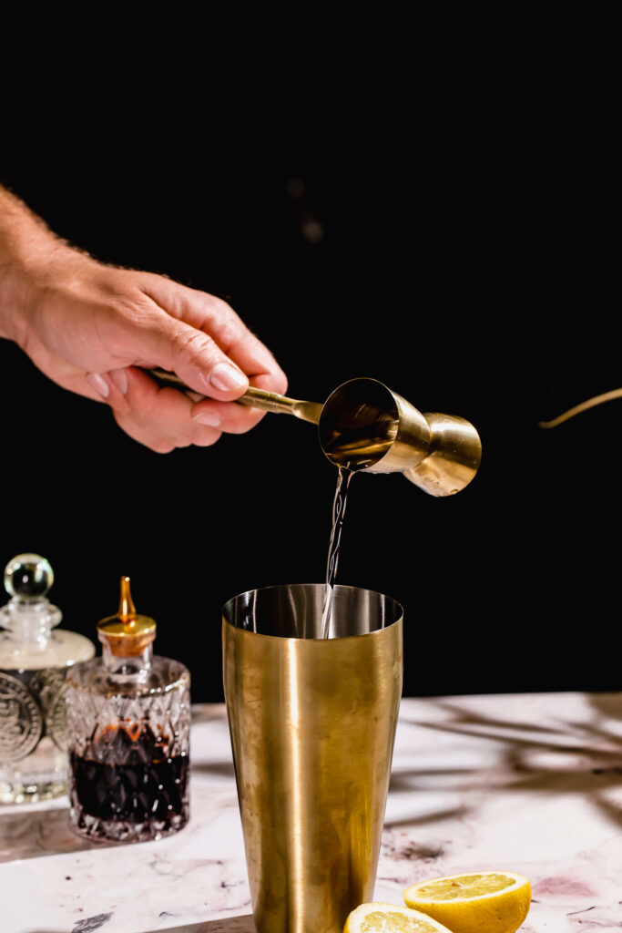 A hand pours gin from a jigger into a gold cocktail shaker surrounded by a bottle, decanter, and lemon halves.