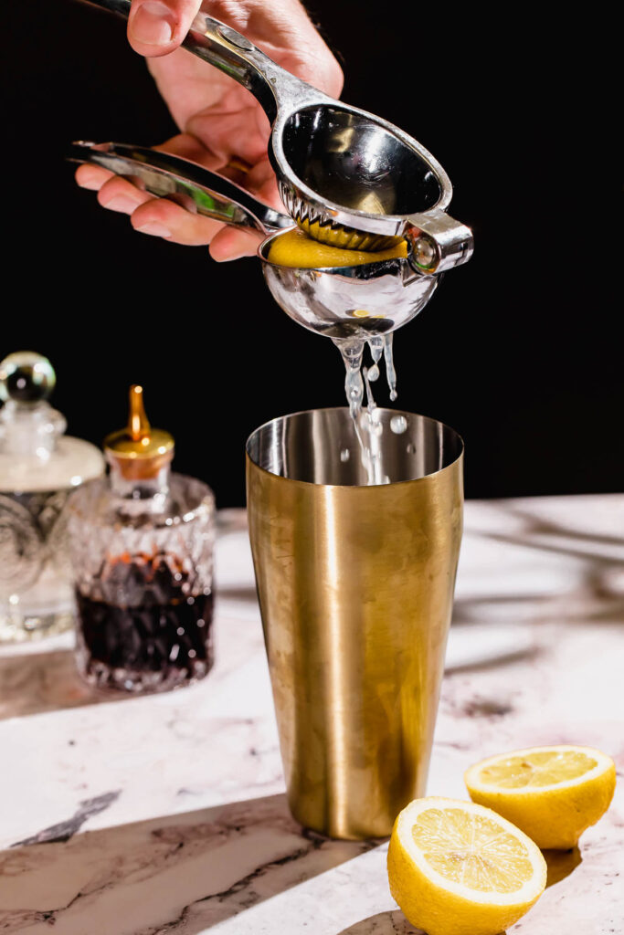 A hand uses a metal juicer to squeeze a lemon half over a brass cocktail shaker, with two lemon halves on a marble counter beside glass bottles.