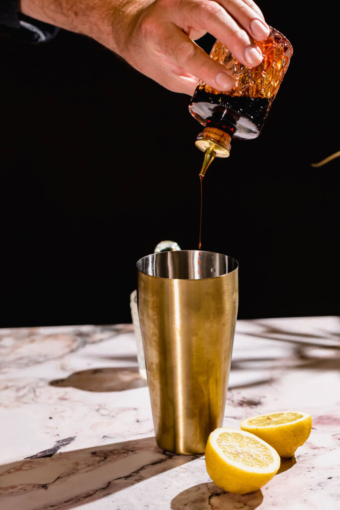 A hand shakes bitters from a small glass bottle into a metal shaker beside two lemon halves on the marble countertop.