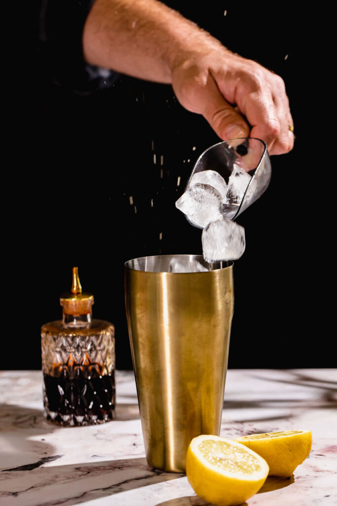 A hand pours ice cubes from a glass into a gold cocktail shaker on a marble surface, with a sliced lemon and spouted bottle nearby.