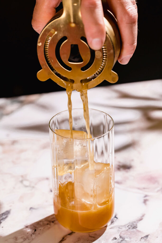 A person strains a cocktail into an ice-filled glass on a marble surface.