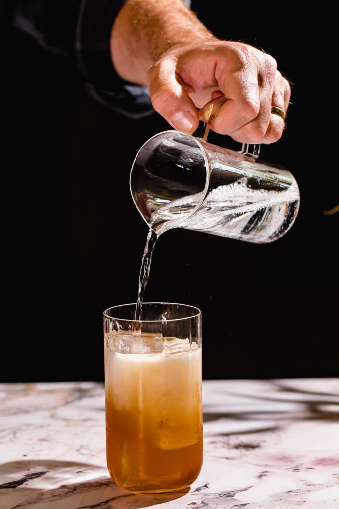 A hand pours tonic from a pitcher into an ice-filled gin sling on a marble surface.