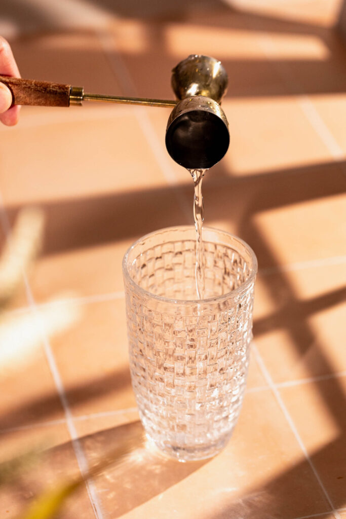 A hand pours vodka from a brass jigger into a textured glass on tiles.