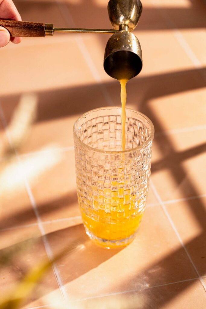 A hand pours pineapple juice from a brass jigger into a textured glass on tiles.