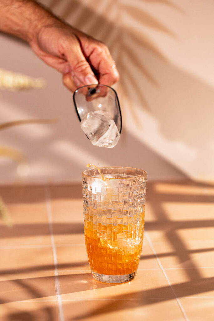 A hand adds ice cubes to a textured glass of orange liquid on a tiled surface.