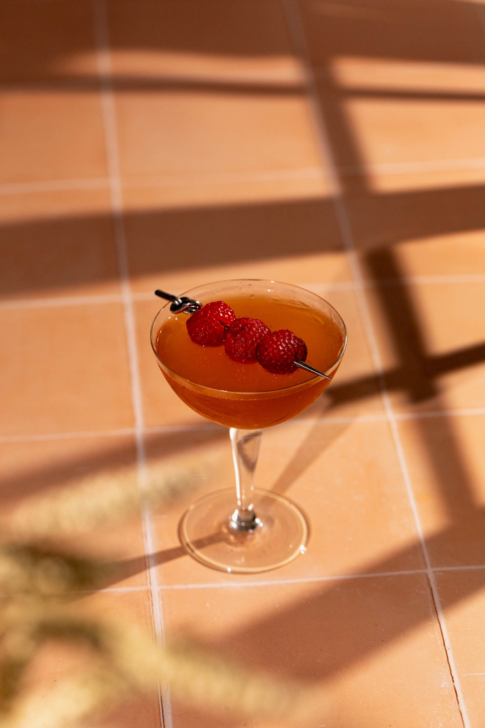 A cocktail glass filled with a French Martini drink, garnished with skewered raspberries, sits on a tiled surface where window frame shadows fall.