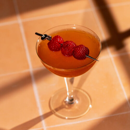 A French Martini cocktail garnished with three skewered raspberries sits on a tiled surface with shadows.