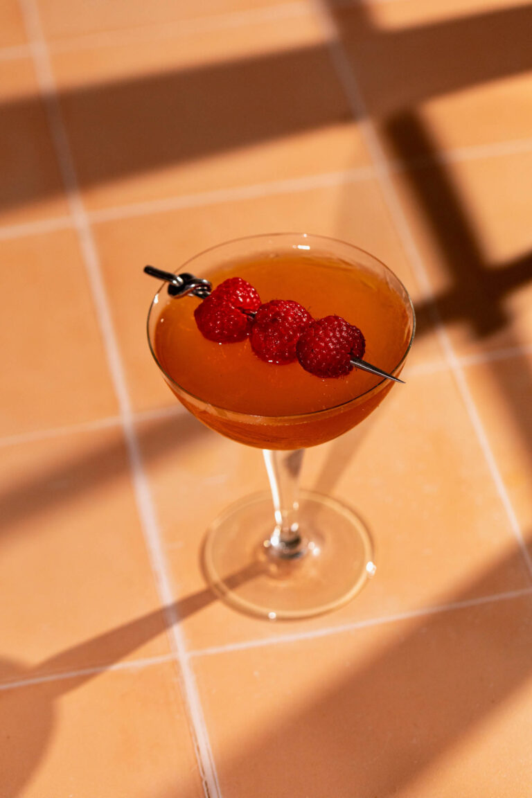 A French Martini cocktail garnished with three skewered raspberries sits on a tiled surface with shadows.