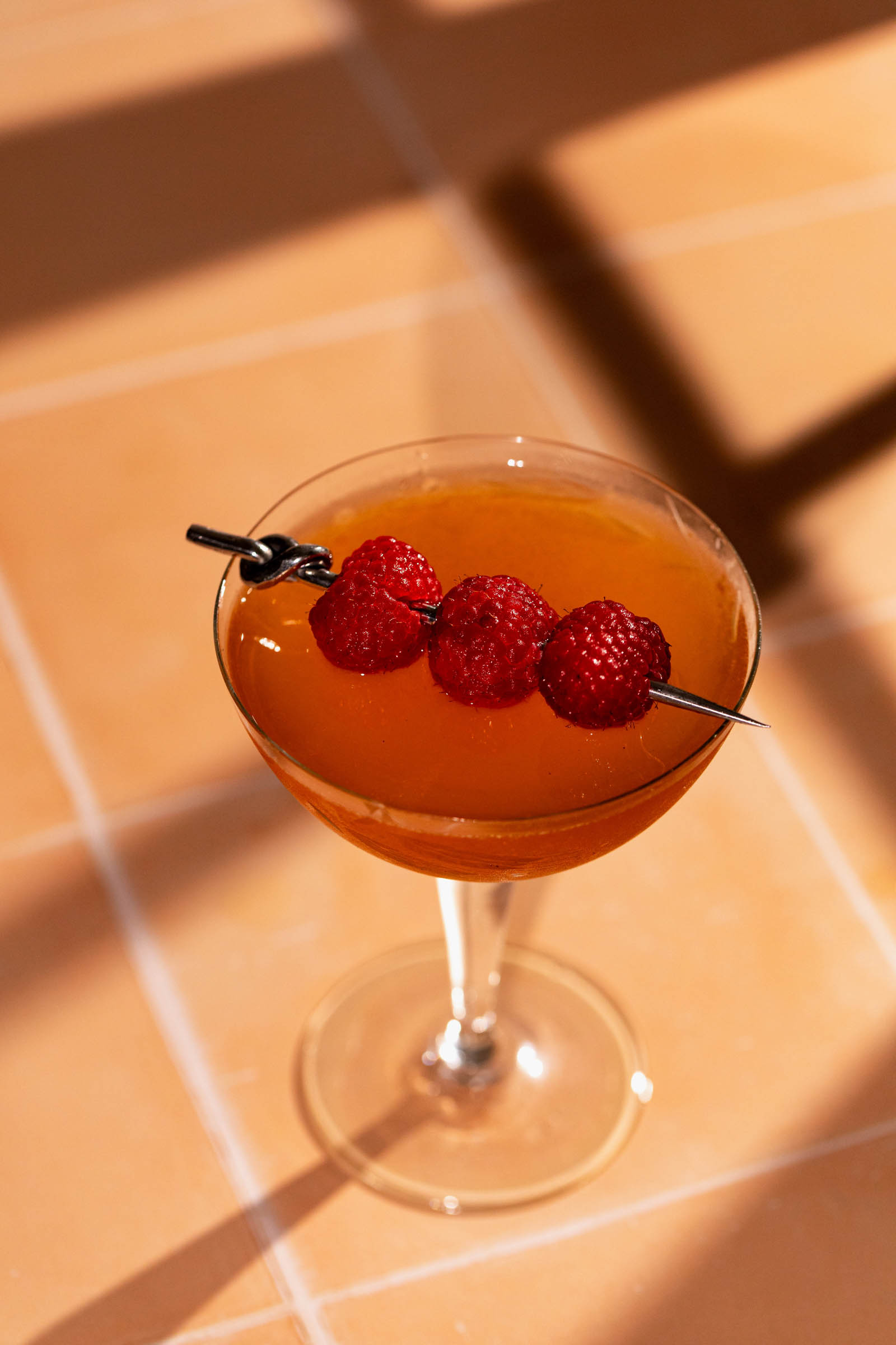 A French Martini cocktail with a skewer of raspberries sits on a sunlit tiled surface.