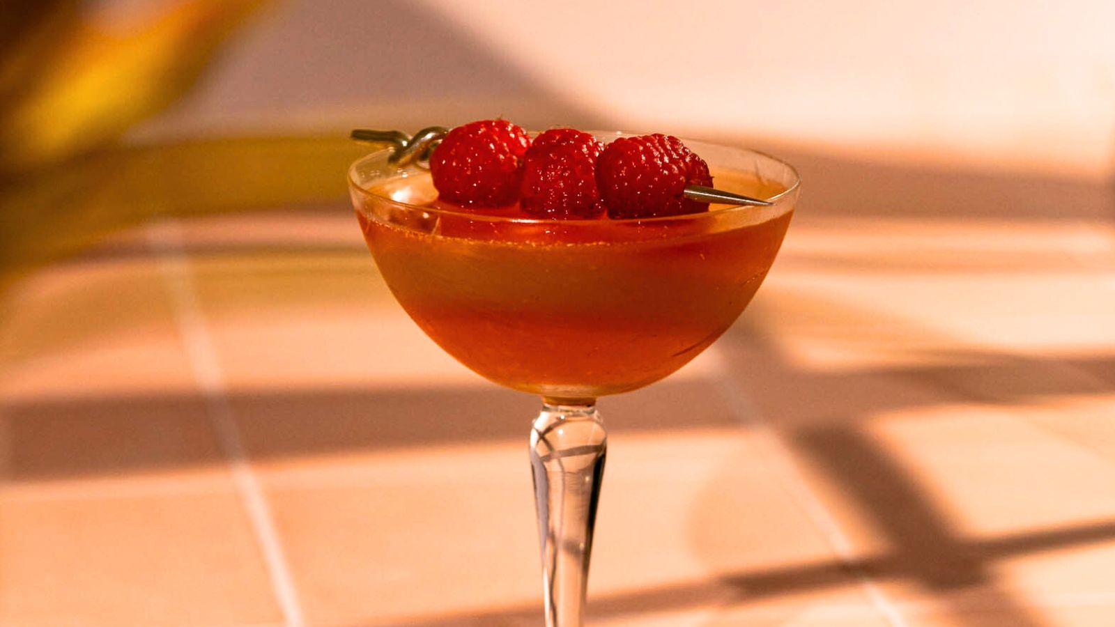 A cocktail in a coupe glass garnished with skewered raspberries casts striped shadows on a tiled surface.