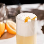 A frothy gin fizz cocktail garnished with a lemon peel sits on a white surface with sliced lemons in the background.