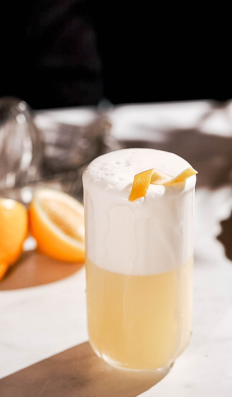 A frothy gin fizz cocktail garnished with a lemon peel sits on a white surface with sliced lemons in the background.