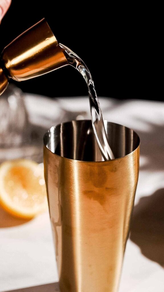 A metal jigger pours gin into a gold cocktail shaker with a lemon slice nearby.