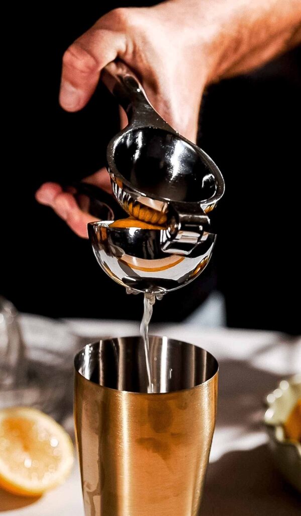 A hand uses a metal citrus press to squeeze juice from a halved lemon into a gold-colored shaker.