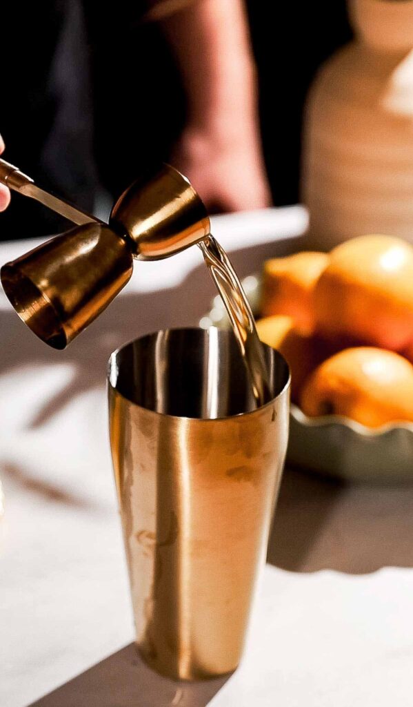 A person pours simple syrup from a jigger into a metal cocktail shaker, with a bowl of oranges nearby.