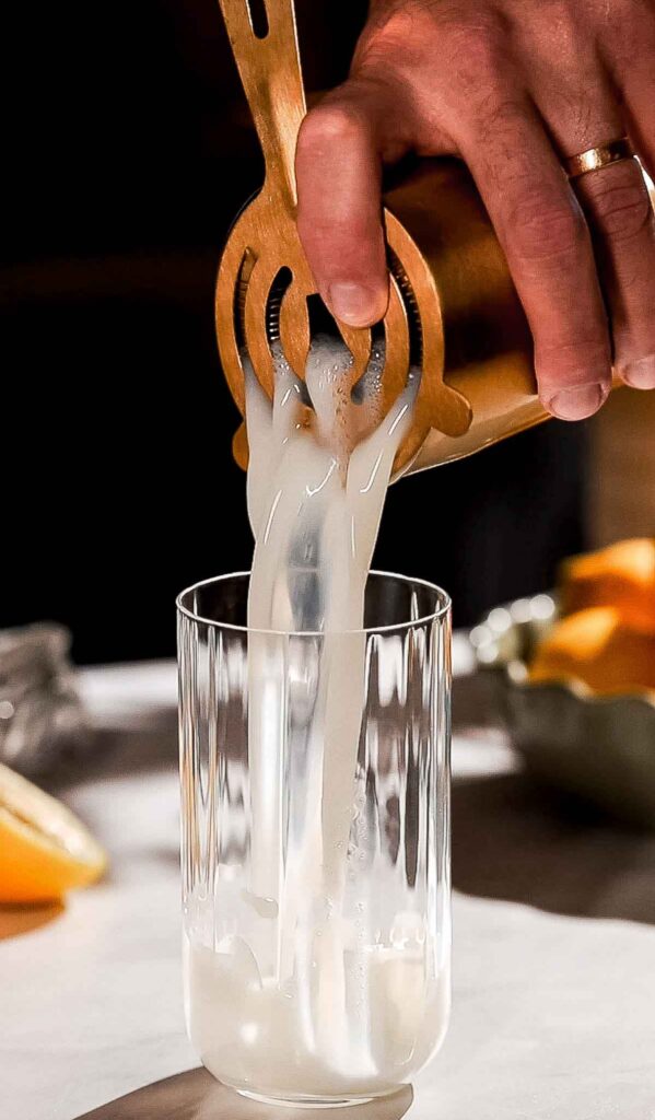 A hand pours a gin fizz through a strainer into a glass, with citrus slices in the background.