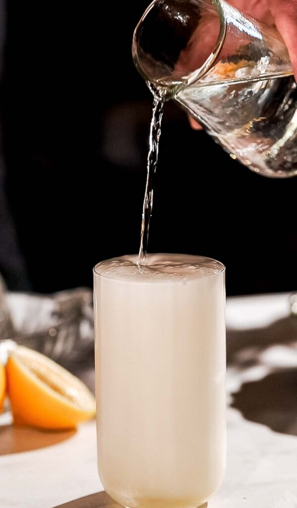 A glass of gin fizz on a table is being topped off with soda water poured from a pitcher, surrounded by lemon wedges.