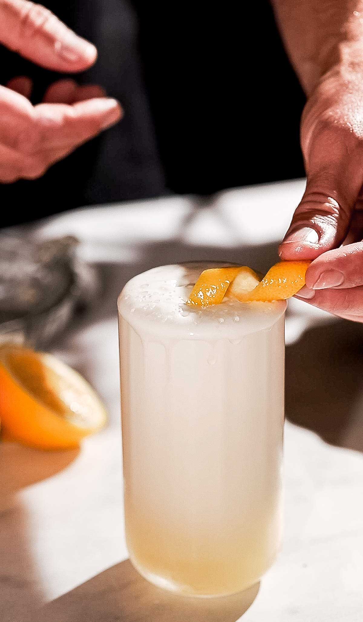 A frothy gin fizz cocktail is garnished with a lemon twist on a marble surface.