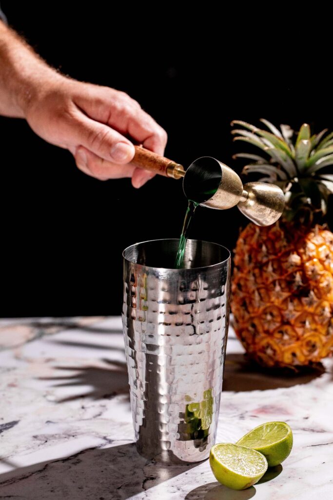 On a marble surface, a person pours Green Chartreuse from a brass jigger into a metal cocktail shaker, with pineapple and lime halves nearby.