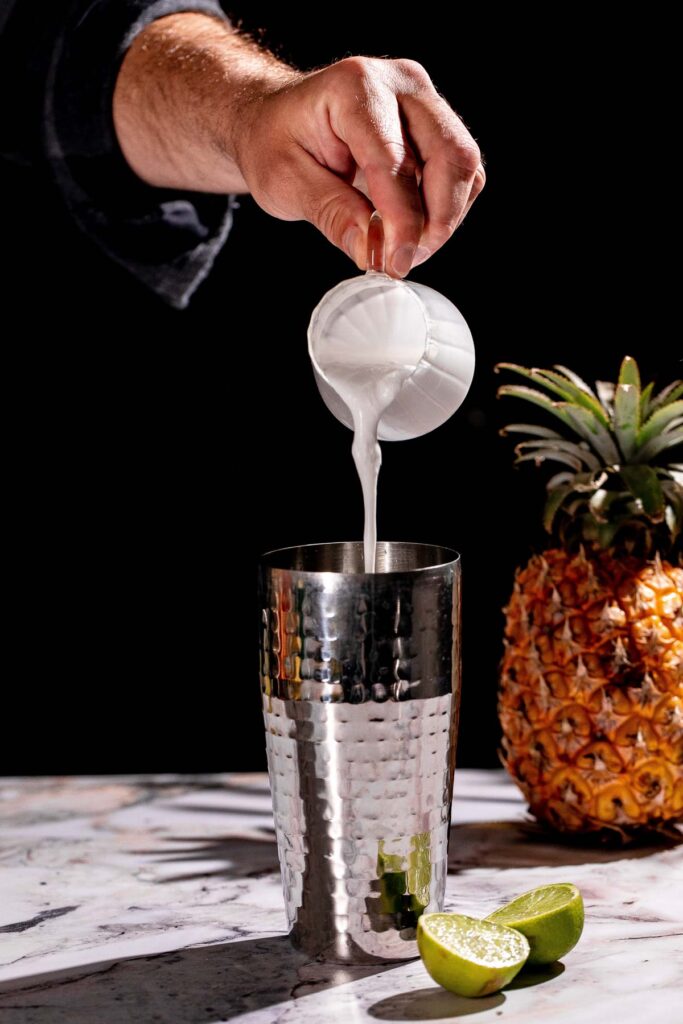 A person pours cream of coconut into a metal shaker beside a pineapple and lime halves on marble.