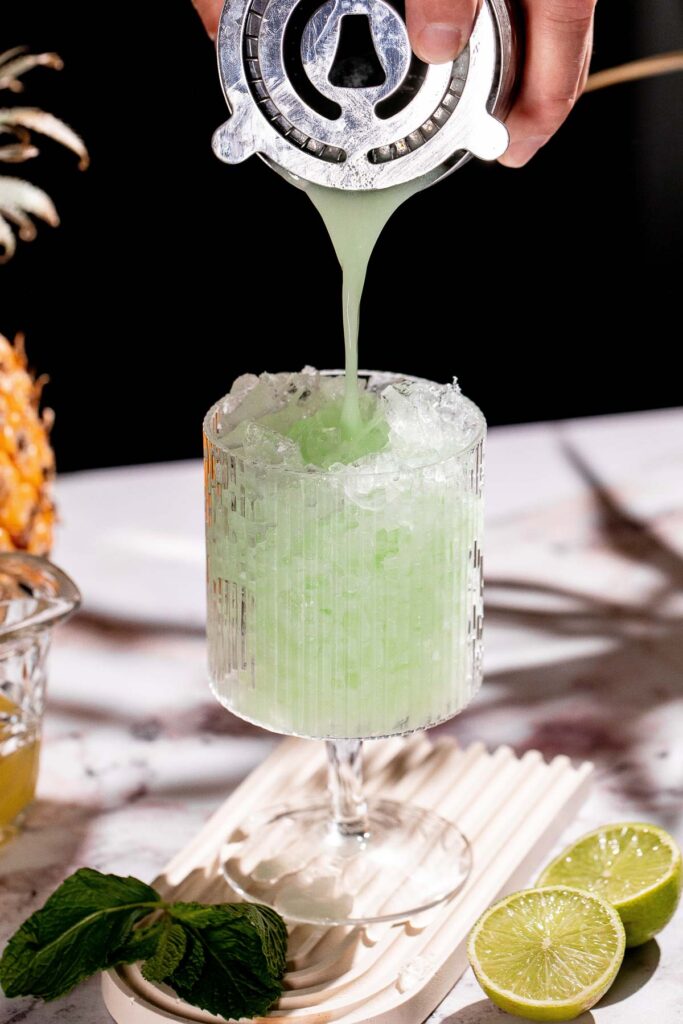 A Piña Verde cocktail is strained into an ice-filled glass surrounded by lime halves and mint leaves.