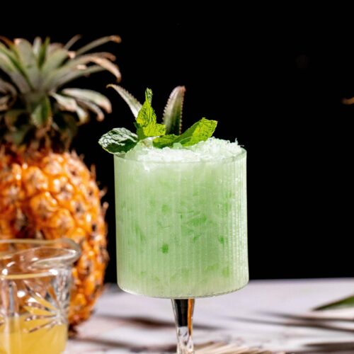 A piña verde cocktail garnished with mint leaves sits in a glass on a table with pineapple, lime, and a small glass of juice against a dark background.