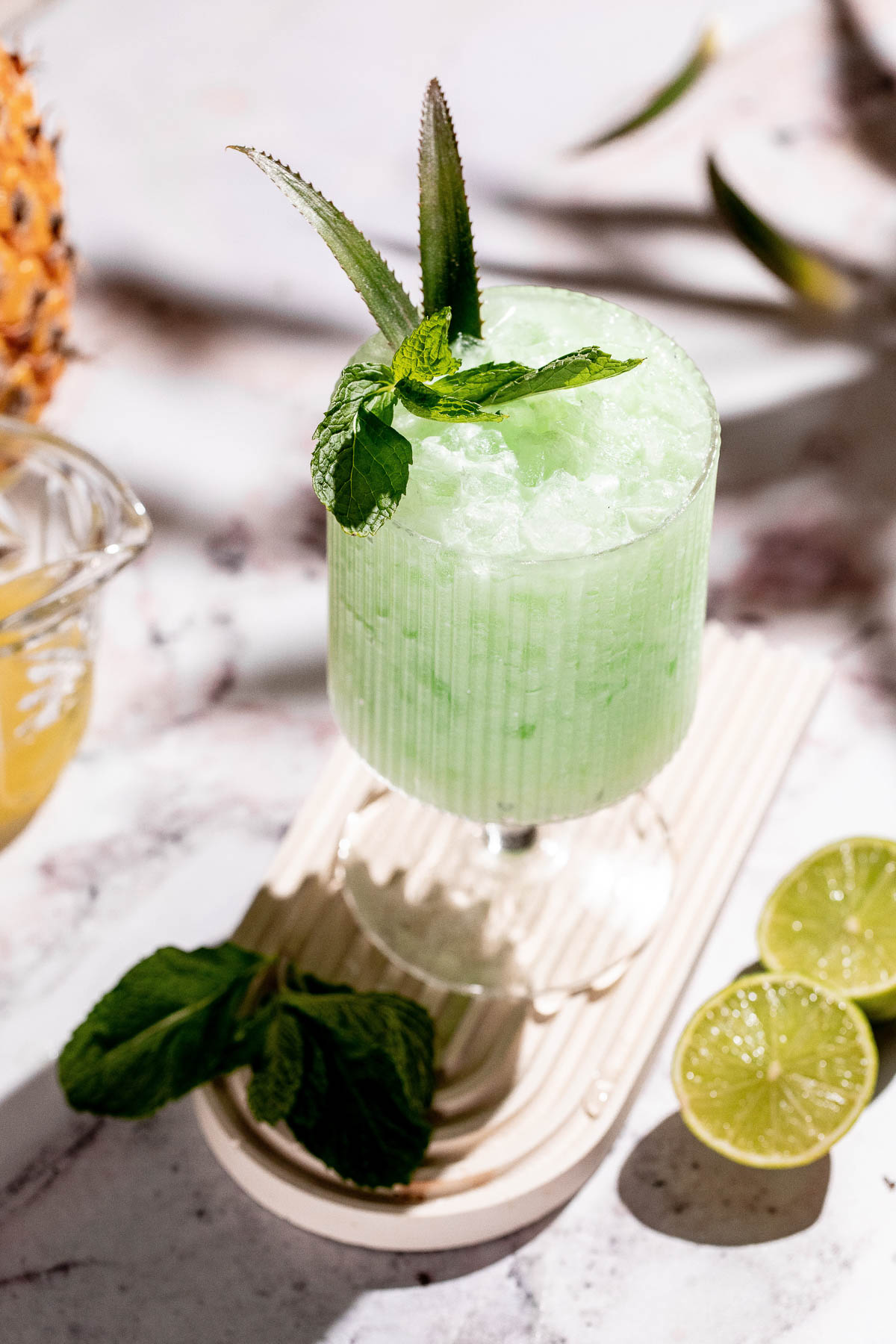 A green icy piña verde in a ridged glass, garnished with mint and aloe, sits on a wooden board with lime halves and mint leaves nearby on a marble surface.
