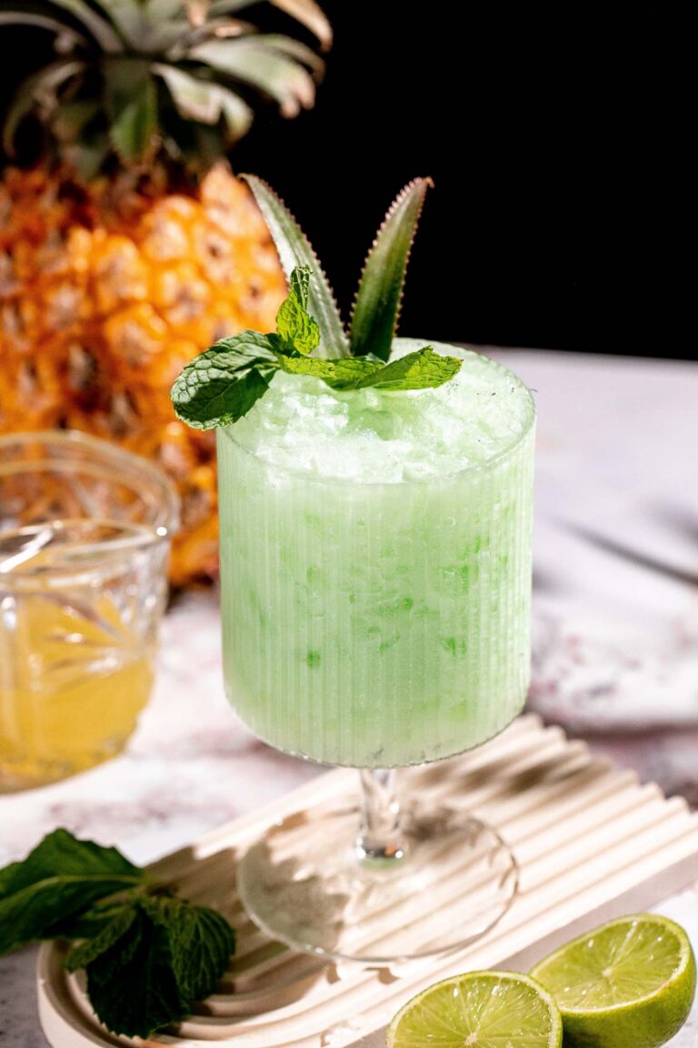 A ribbed glass filled with a piña verde cocktail garnished with mint and pineapple leaves sits alongside lime slices, with a pineapple and a glass of yellow liquid in the background.