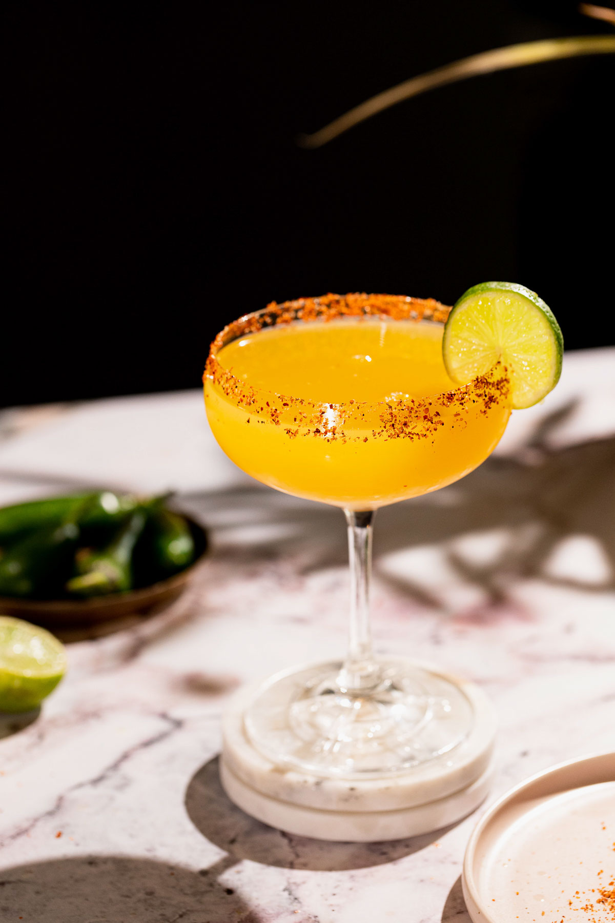 A spicy mango margarita cocktail with a chili-salt rim and lime slice sits in a stemmed glass, with green peppers on a marble surface in the blurred background.
