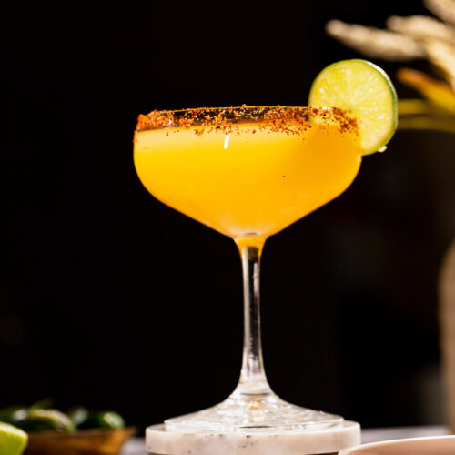 A spicy mango margarita with a chili-rimmed edge and lime slice sits on coasters.