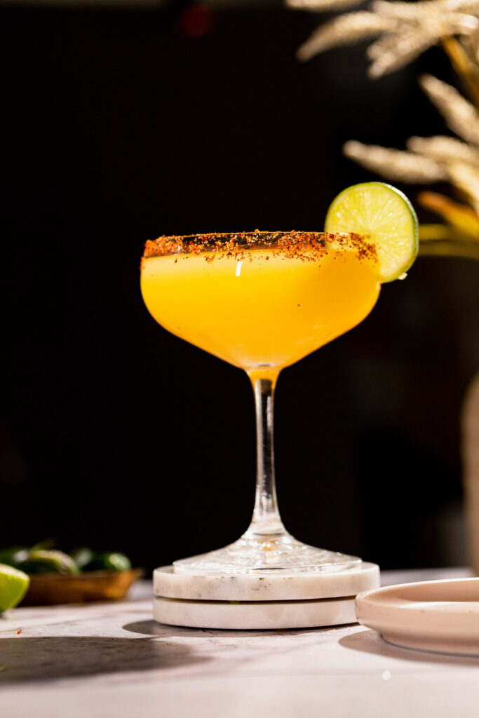 A spicy mango margarita with a chili-rimmed edge and lime slice sits on coasters.