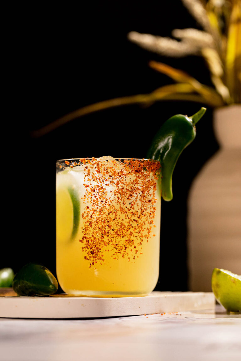 A spicy margarita with ice, garnished with jalapeño and lime slices, has a chili powder rim and a nearby lime wedge.