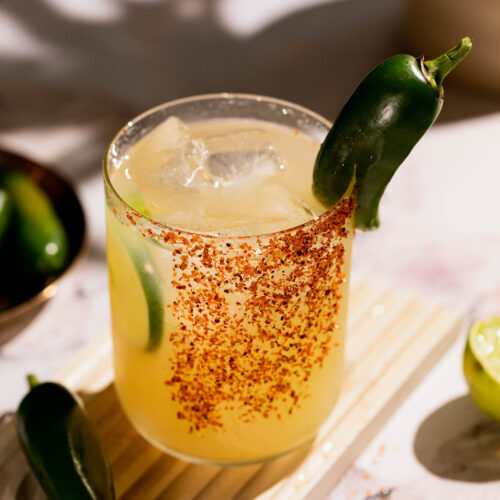 A spicy margarita cocktail with ice is garnished with a jalapeño, lime wedge, and a chili powder-coated rim, alongside a whole jalapeño.