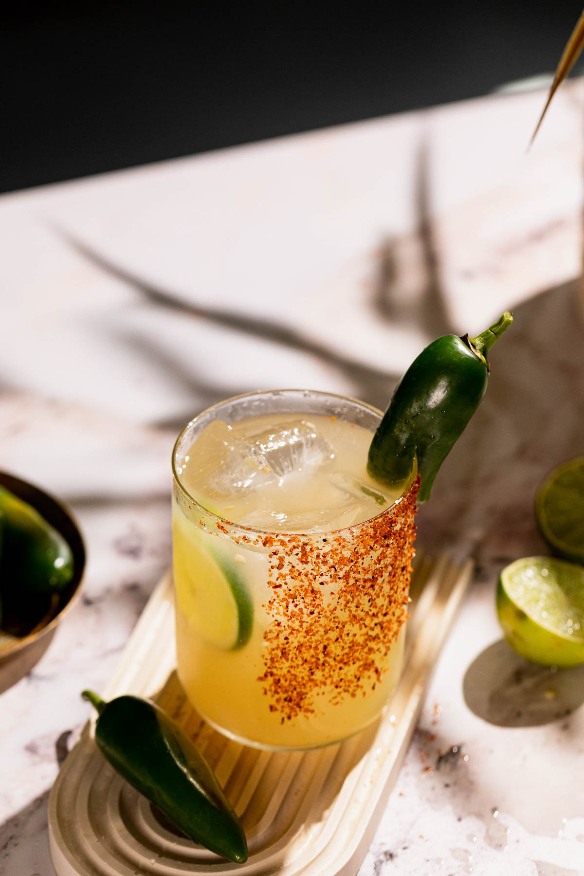 A spicy margarita with a chili-powder rim, garnished with lime and jalapeño, is served on ice in a glass on a marble surface with nearby limes and jalapeños.