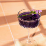 A purple cocktail in a crystal coupe glass, garnished with a small purple flower, sits on a tiled surface in natural light.