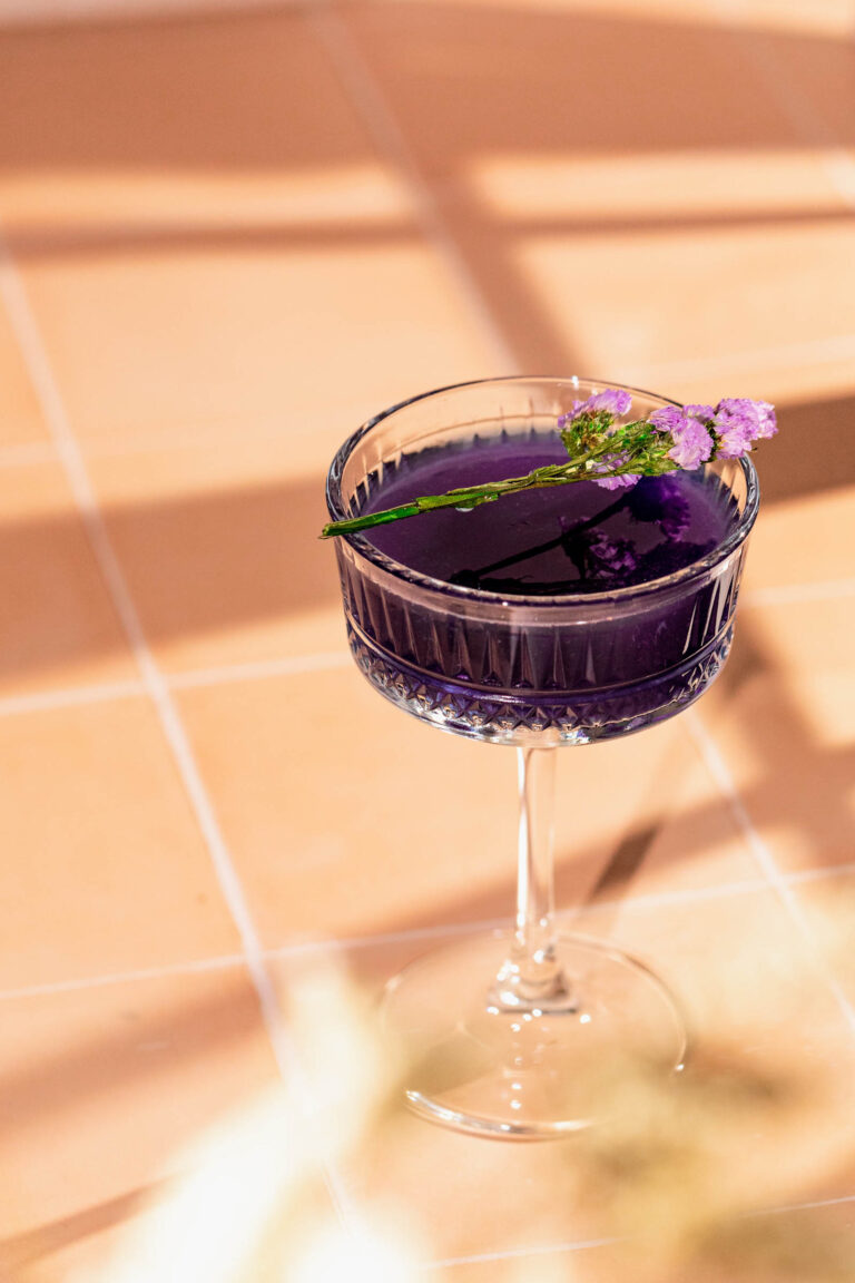 A purple cocktail in a crystal coupe glass, garnished with a small purple flower, sits on a tiled surface in natural light.