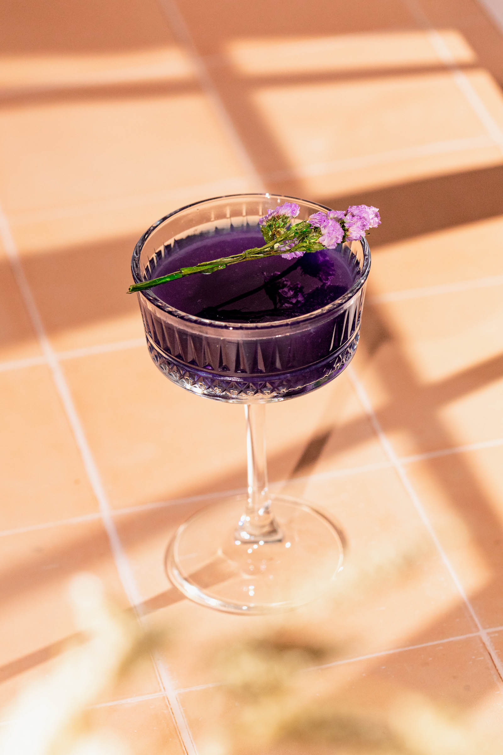 A purple cocktail in a decorative glass on a tile surface is garnished with small purple flowers and a green stem.