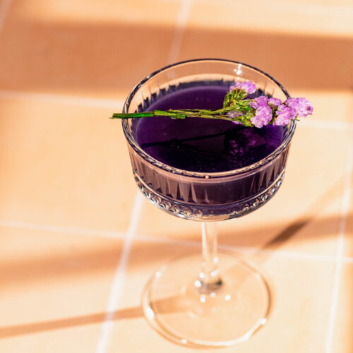 A purple cocktail in a clear glass, topped with a small sprig of purple flowers, sits on a light tiled surface.