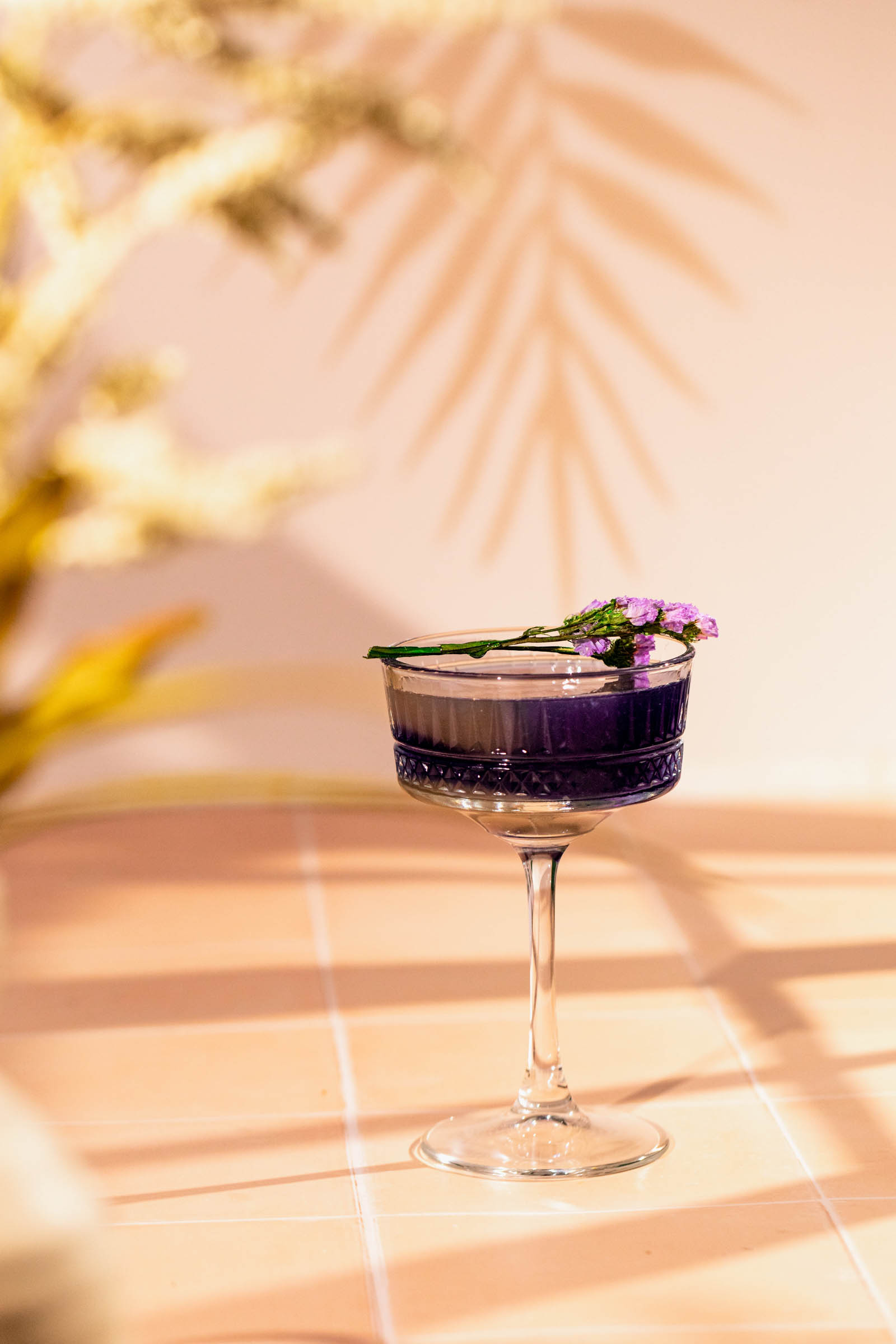 A purple cocktail adorned with small flowers sits on a tiled surface, with a plant casting a shadow in the background.