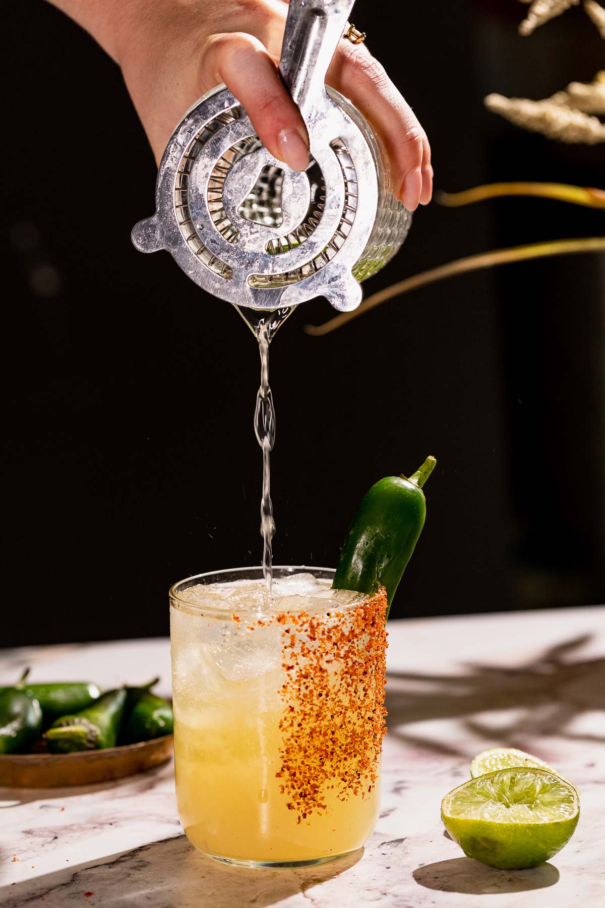 A margarita is strained into a salted-rim glass garnished with jalapeño, set on a marble surface surrounded by limes and jalapeños.