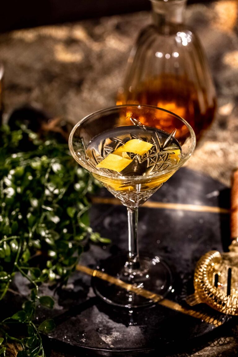 A cocktail glass with a clear 50/50 martini and lemon twist sits on a dark surface, with a decanter and green leaves in the background.