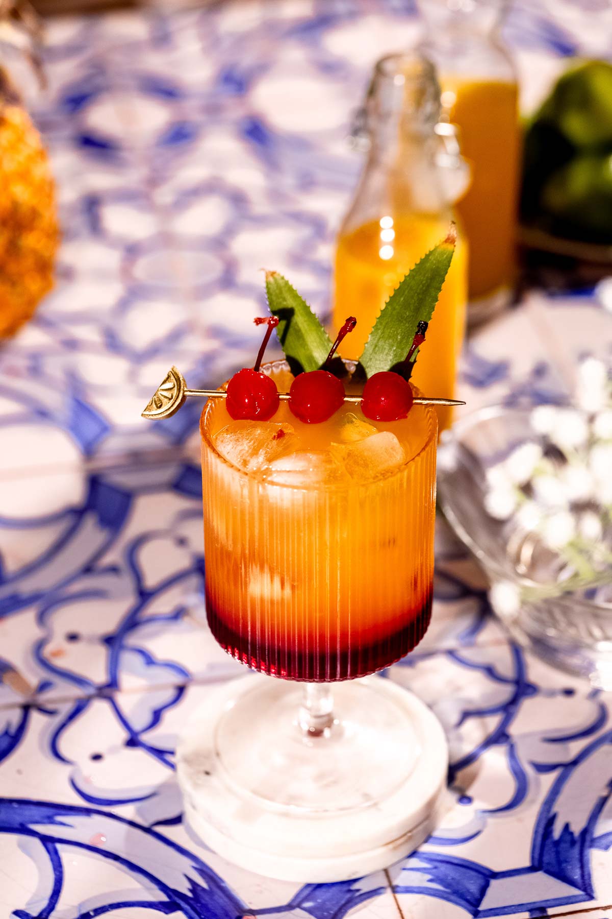 A Bahama mama garnished with pineapple leaves and skewered cherries sits on a blue-patterned surface, with a blurred bottle and fruit in the background.