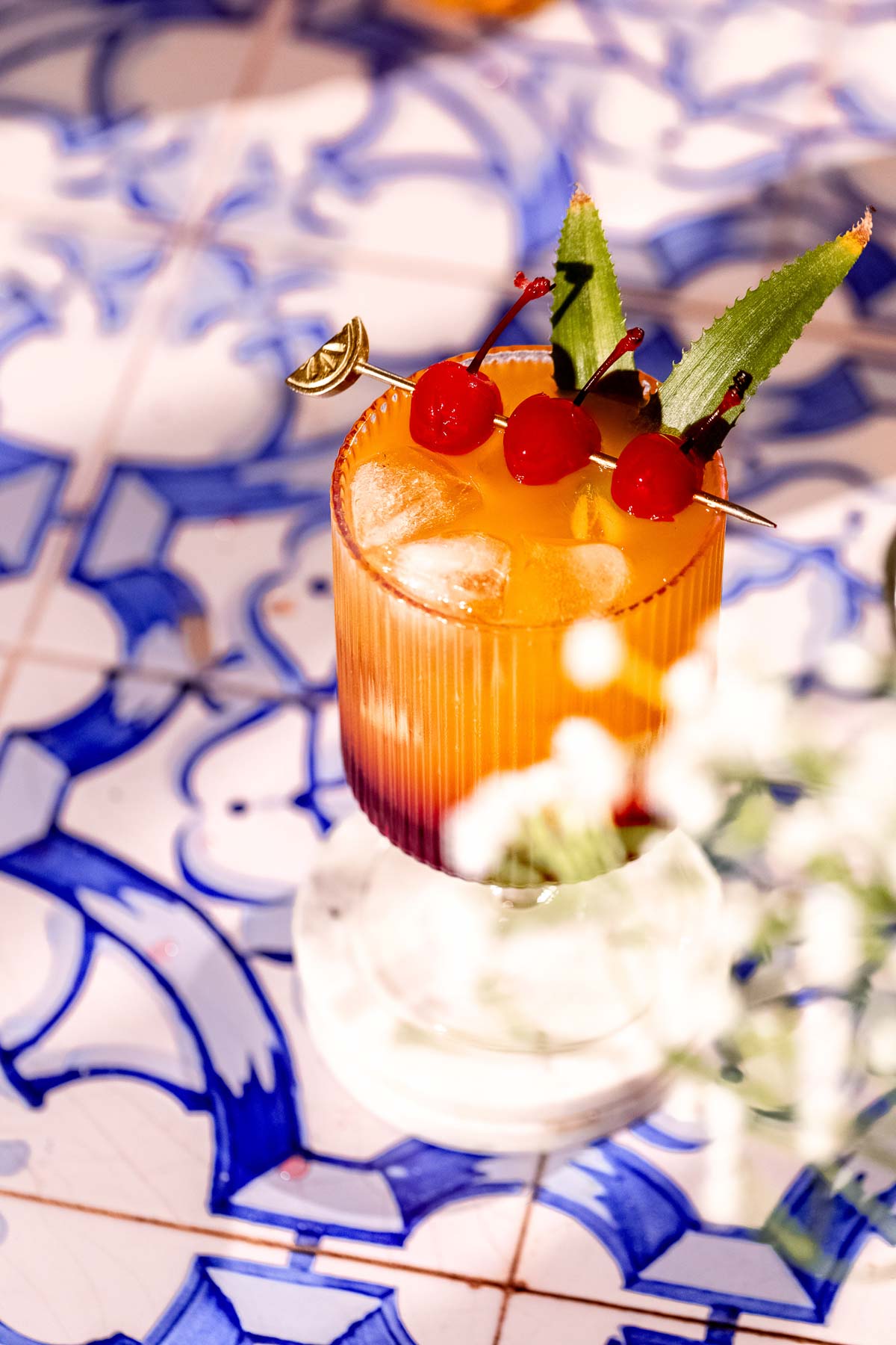 A Bahama mama cocktail with ice, cherries, and leaves sits on a blue and white patterned tile table.