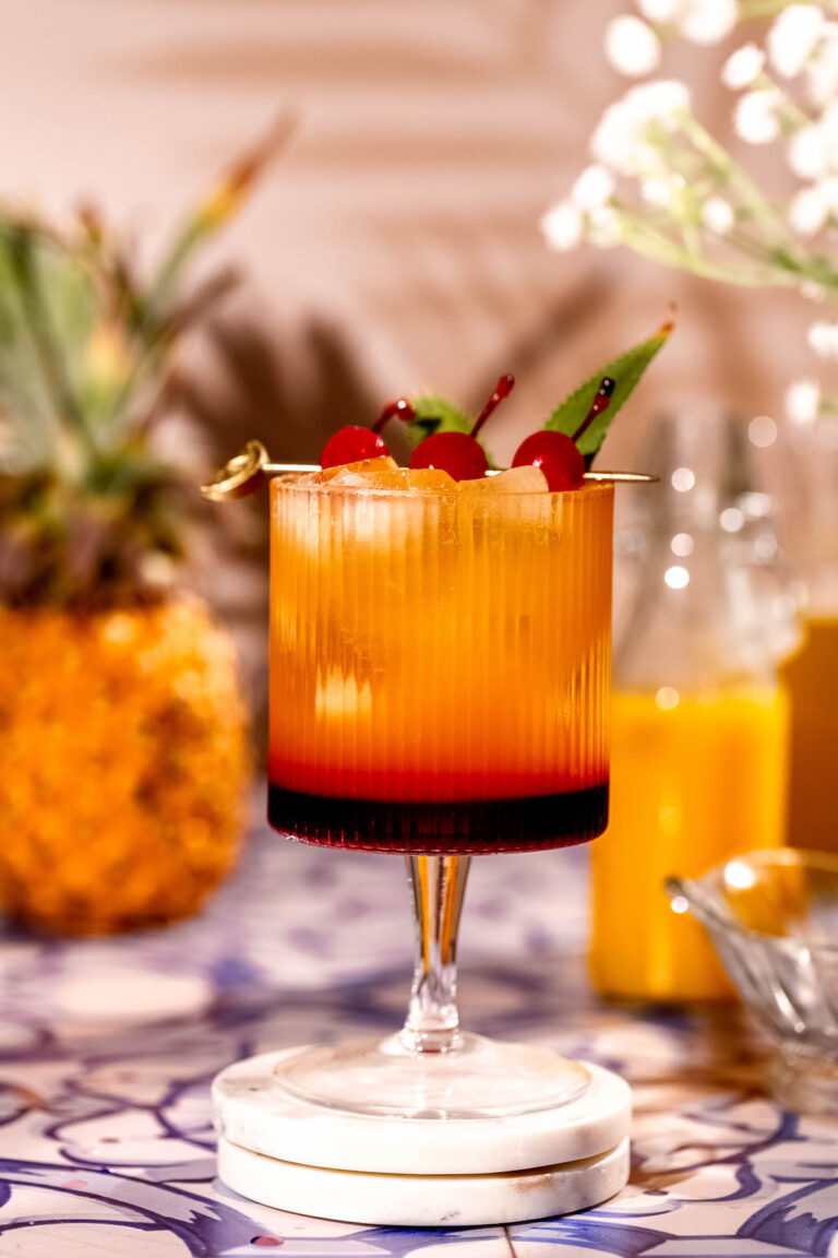 A layered orange and red Bahama mama cocktail garnished with cherries and pineapple leaves sits on a patterned surface, with a whole pineapple and bottle in the background.