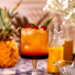A Bahama Mama cocktail garnished with cherries and mint sits on a tiled surface with blurred pineapple and orange juice bottles in the background.