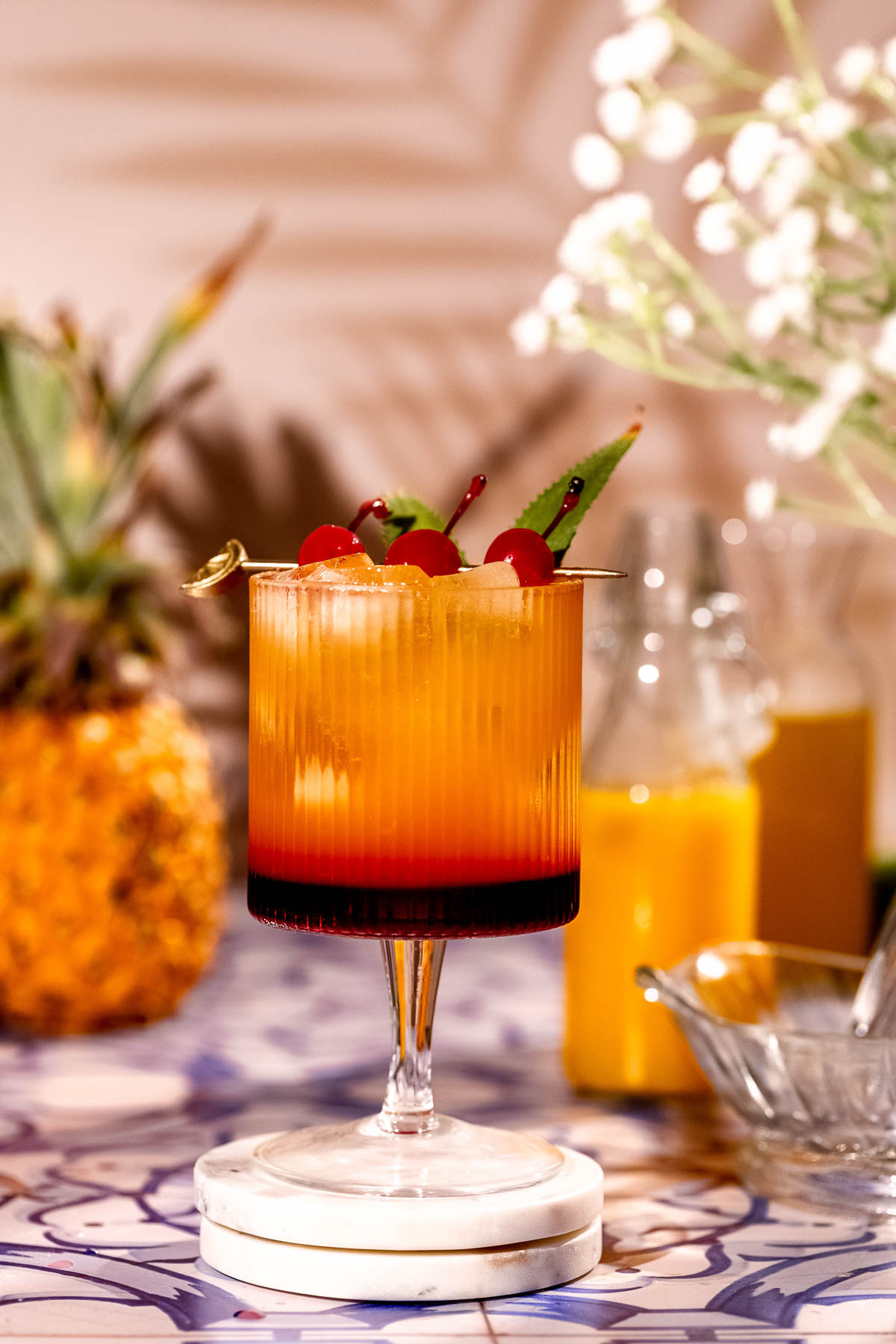 A Bahama Mama cocktail garnished with cherries and mint sits on a tiled surface with blurred pineapple and orange juice bottles in the background.