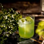 A gin basil smash cocktail with leaf garnish rests on a dark slate amidst greenery.